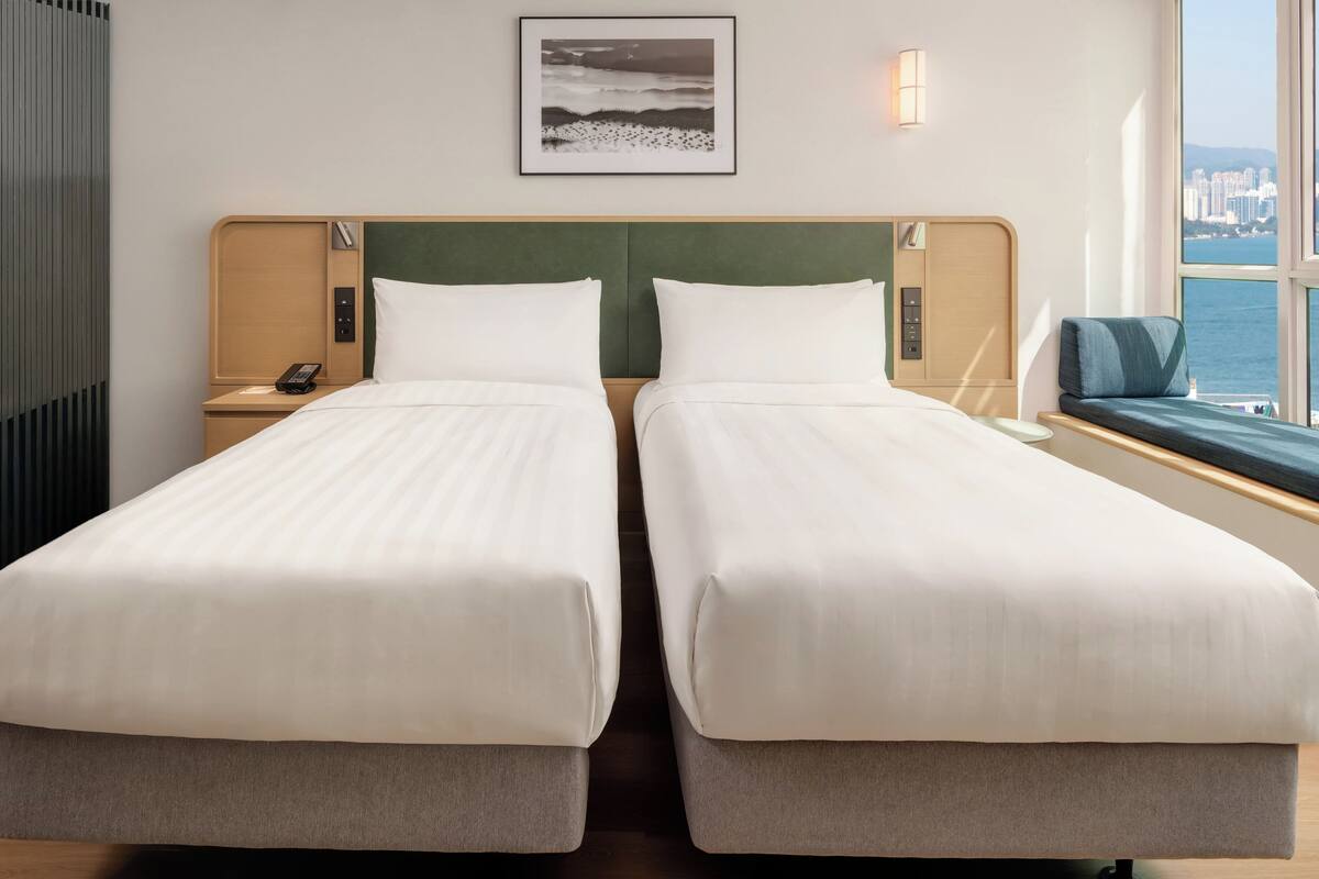 A hotel room with two beds, a view of the sea, a framed picture on the wall, and a nightstand with a phone.