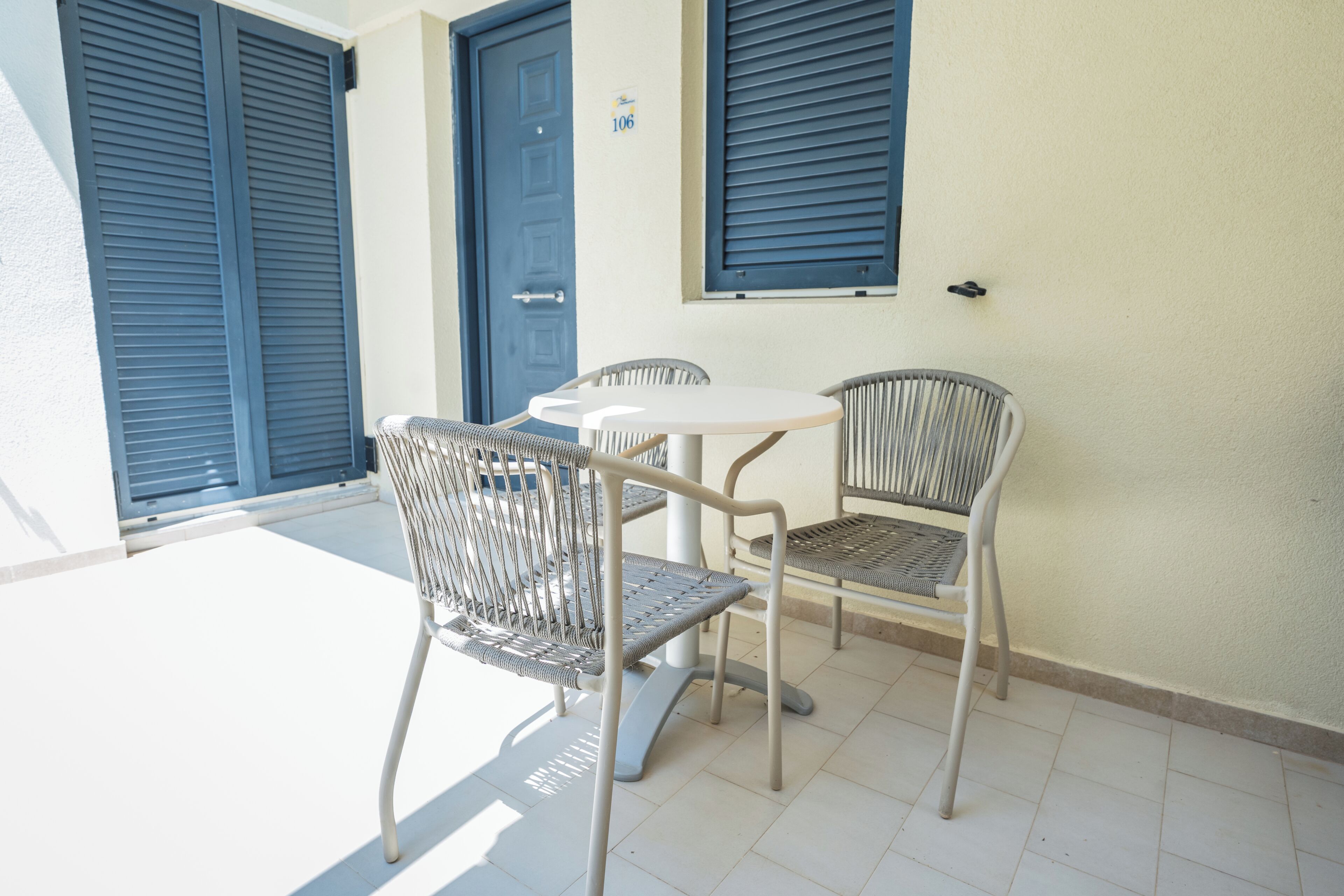 A patio area with a round table and two wicker chairs, a blue door, and a window with shutters.
