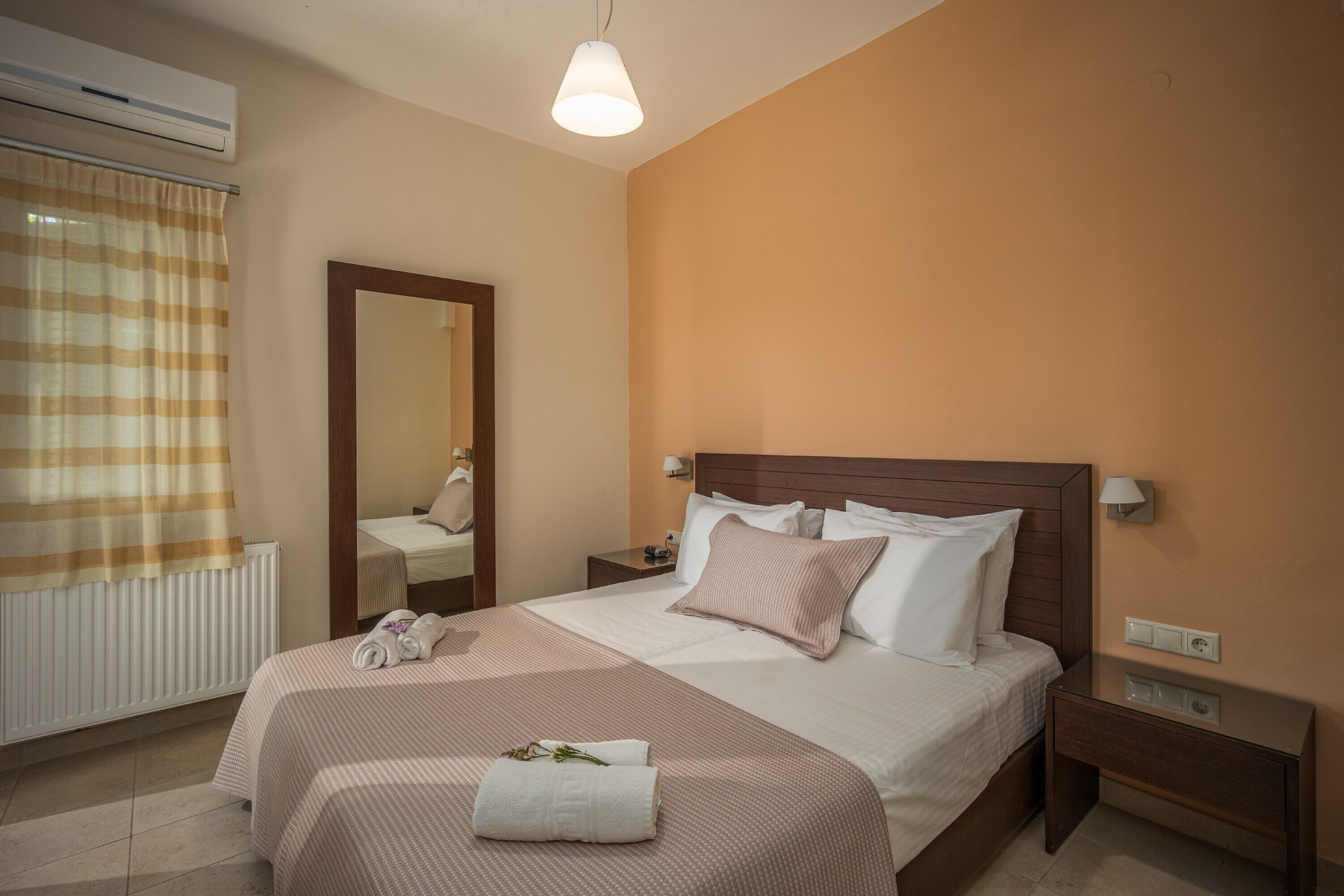 A neatly arranged hotel room with a large bed, a bedside table, a nightstand, a mirror, and an air conditioning unit.