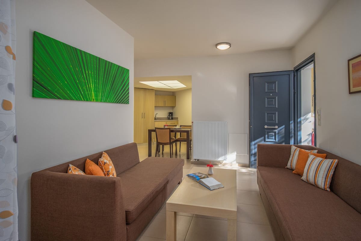 A living room with two brown sofas, a coffee table, and a green abstract painting. There is a kitchen in the background with a dining table and chairs.