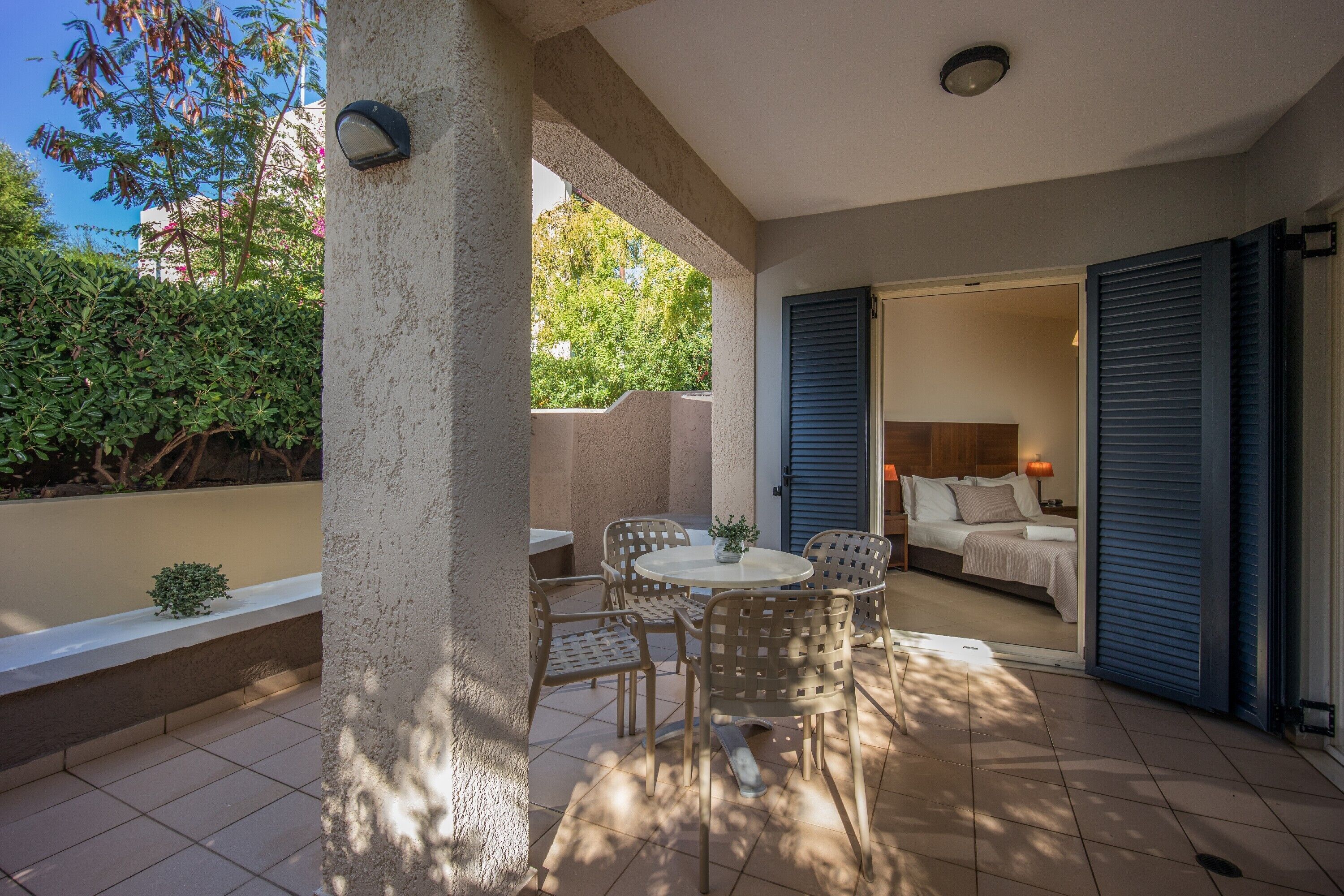 A covered patio with a round table and chairs, a small plant, and a view of a bedroom with a bed and nightstand.