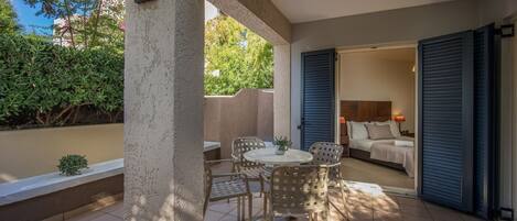 A covered patio with a round table and chairs, a small plant, and a view of a bedroom with a bed and nightstand.