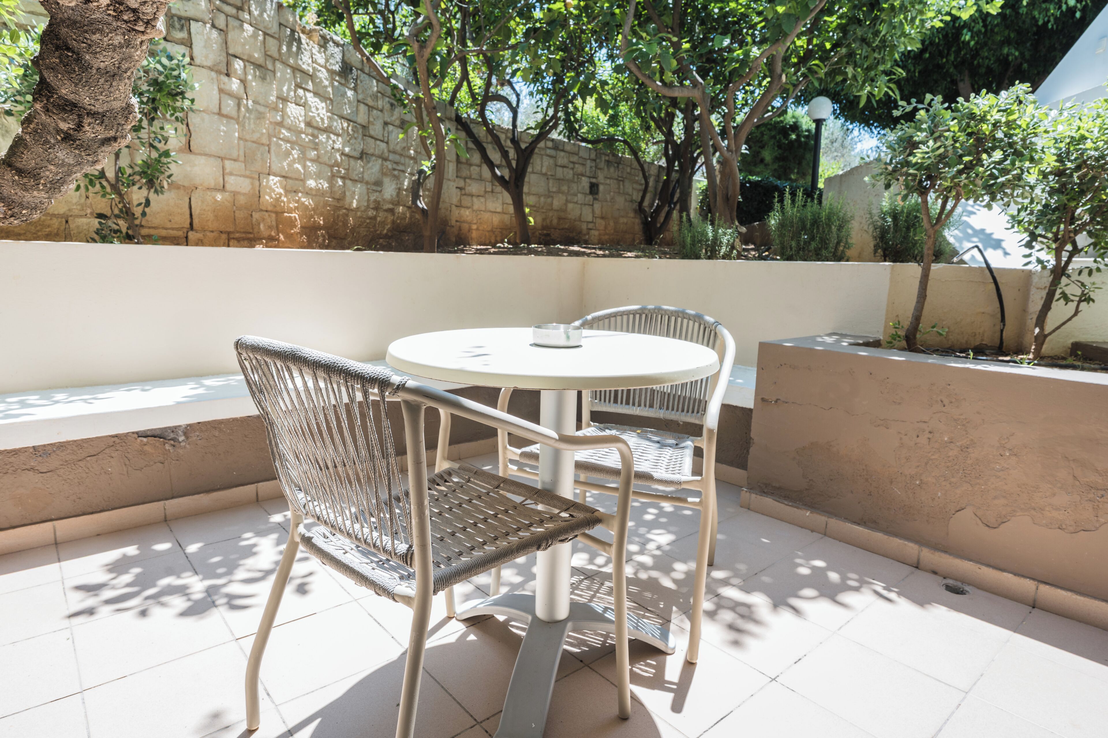 Un patio al aire libre con una mesa redonda y dos sillas de mimbre, un muro de piedra y árboles que dan sombra.