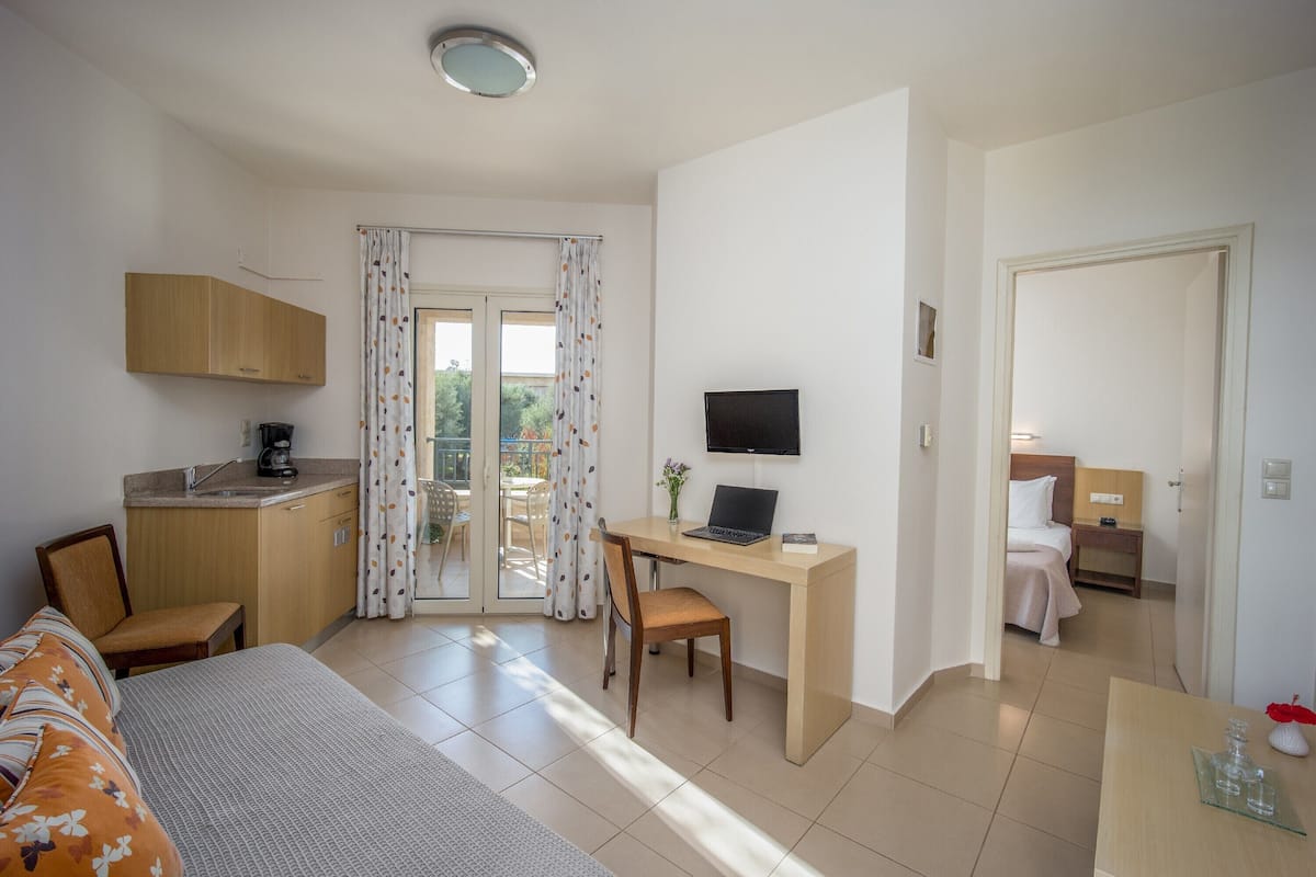 A hotel room with a bed, a desk with a laptop, a TV, a kitchenette, and a balcony with curtains.