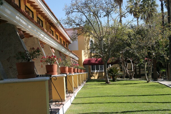 A courtyard with a row of potted plants, a well-maintained lawn, and a building with a tiled roof and yellow walls.