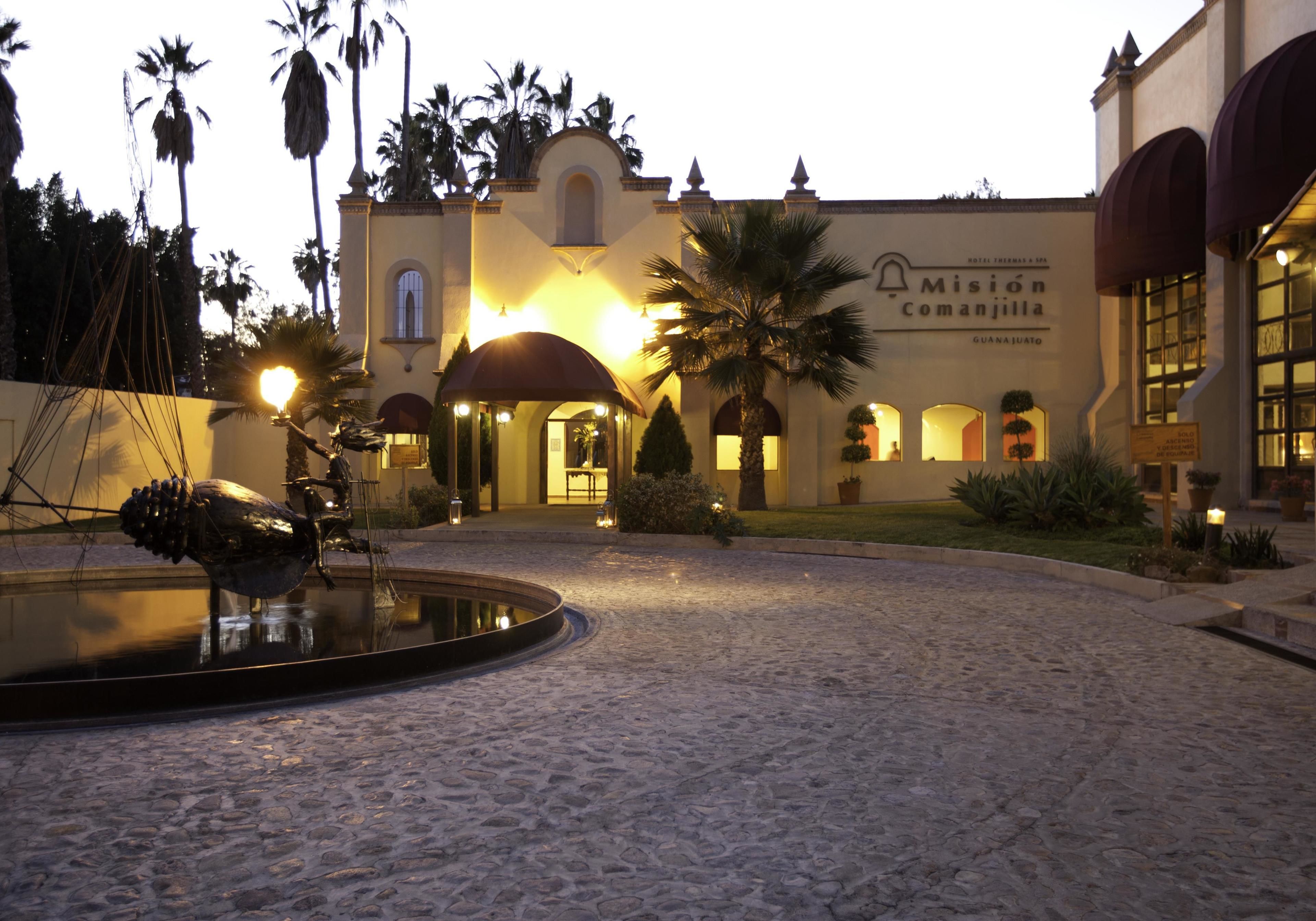 A courtyard with a fountain, palm trees, and a building with a sign reading "Misión Comanjilla."