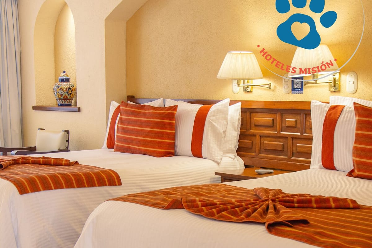Ein Hotelzimmer mit zwei Betten, jeweils mit weißer und orangefarbener Bettwäsche, einem Holzkopfteil und einer Wandlampe.