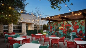 An outdoor patio with tables and chairs, string lights, and a mural of flowers. - Hilton Garden Inn Dallas Richardson (Richardson)
