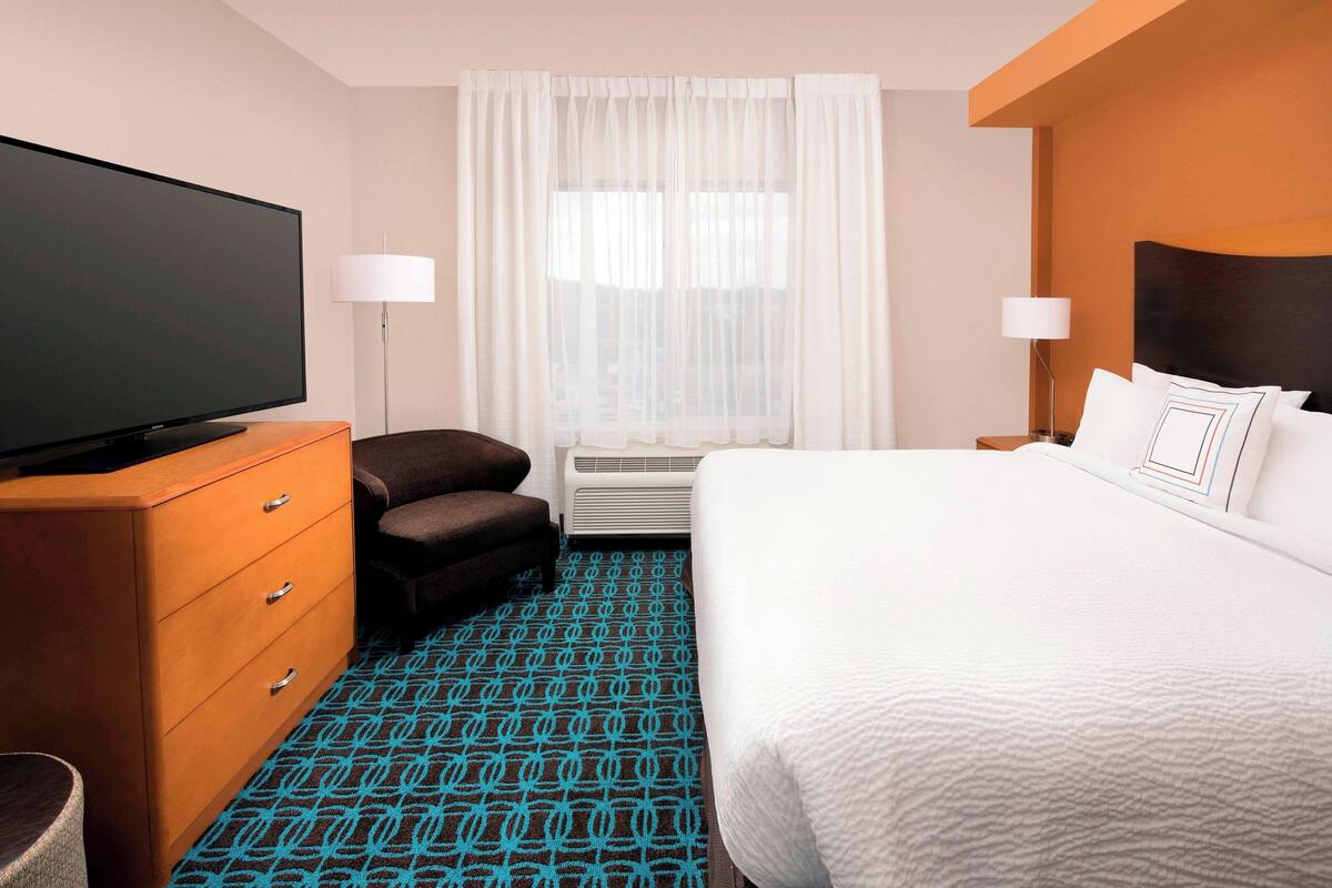A hotel room with a large flat-screen TV, a sofa, a bed with white bedding, and a patterned carpet.