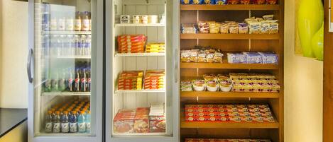 A small grocery store with two refrigerators stocked with beverages and snacks, shelves with packaged goods, and a 'market' sign on the wall.