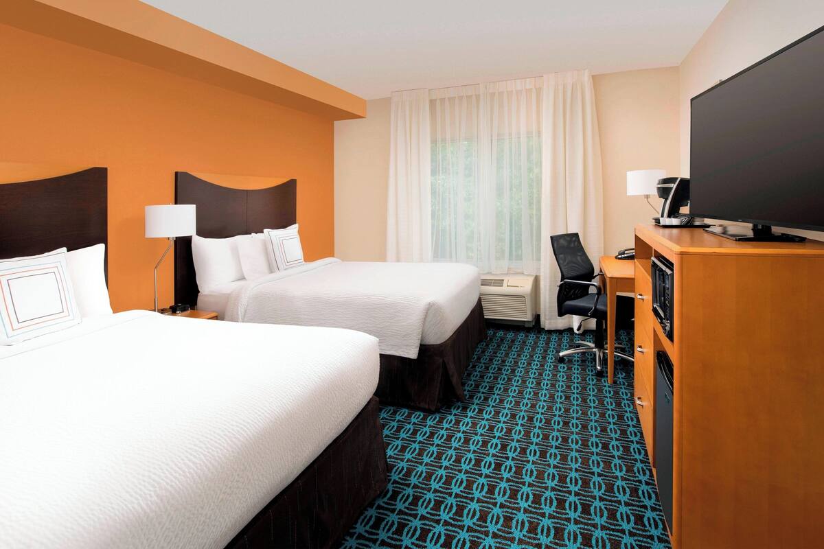 A hotel room with two beds, a desk with a computer, a TV, and a large window with curtains.