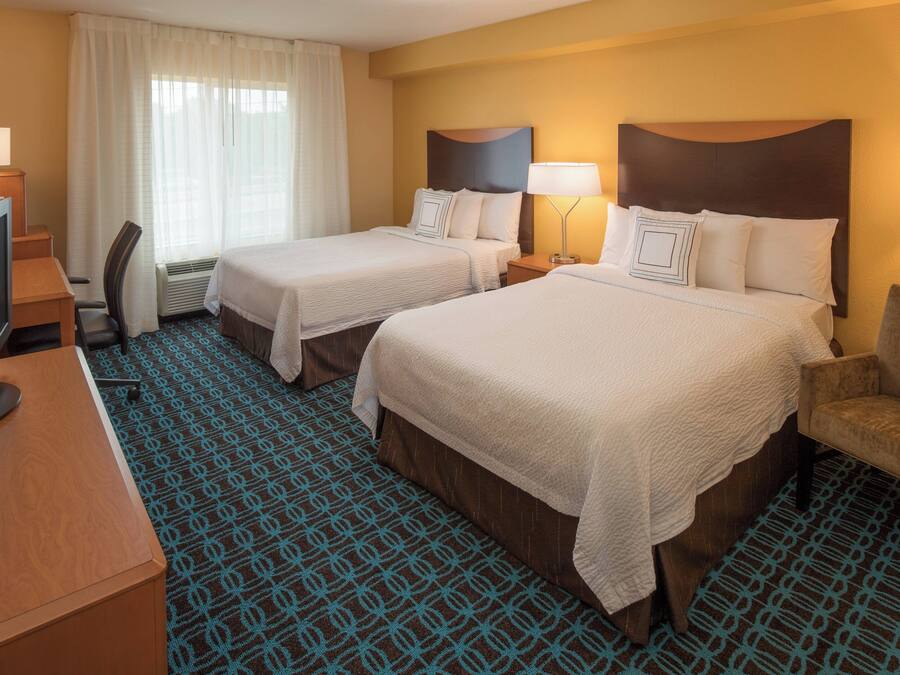 Habitación de hotel con dos camas, un escritorio con televisor, una silla y una alfombra estampada.