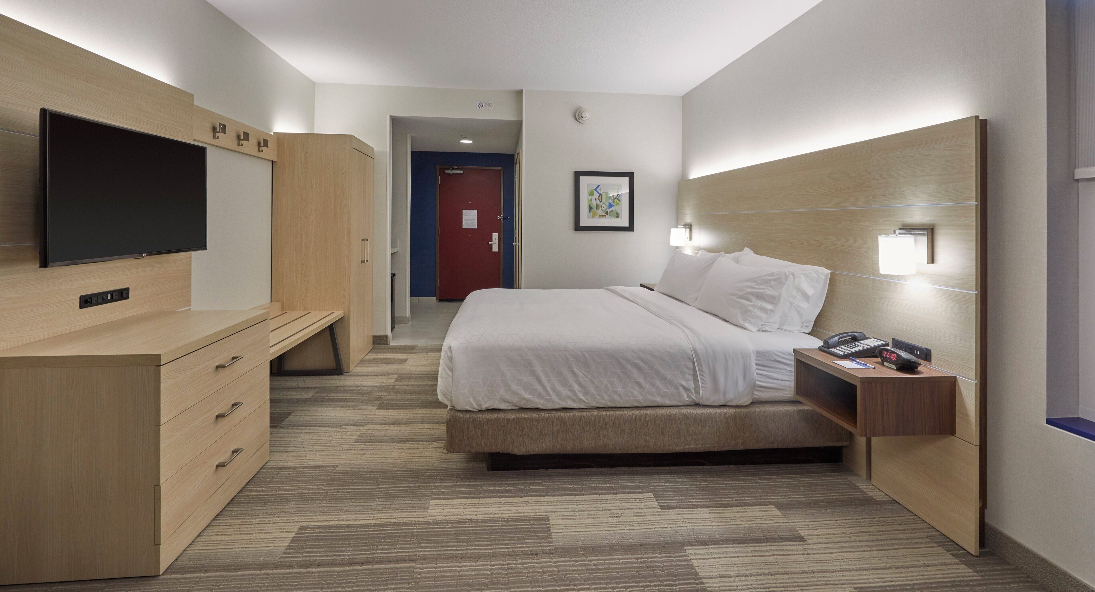 Habitación de hotel con cama, mesita de noche, televisor, armario y una puerta que da a otra habitación.