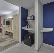 Cocina compacta con microondas, cafetera y refrigerador. En la habitación contigua, hay un escritorio de madera y un televisor.