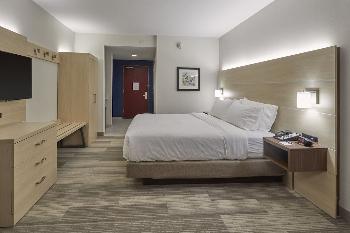 Una habitación de hotel moderna con una cama grande, un televisor, un escritorio con cajones y una mesita de noche con teléfono y lámpara.