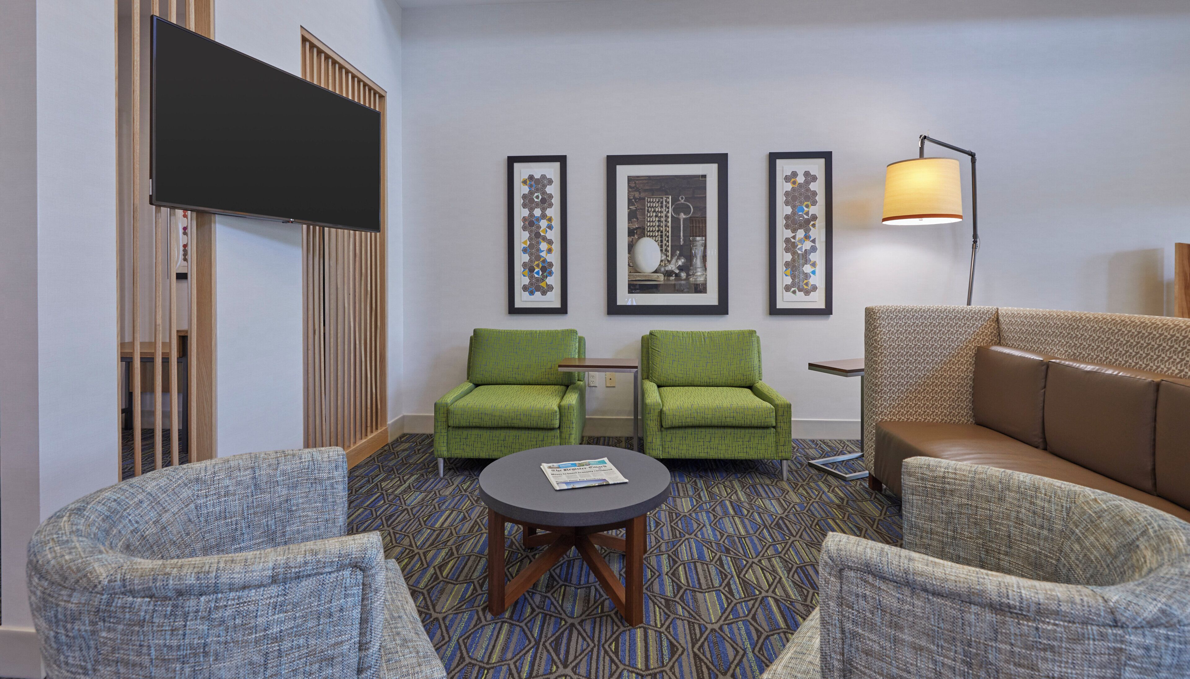 A hotel room with a flat-screen TV, two green armchairs, a patterned sofa, a small round table, and three framed artworks on the wall.