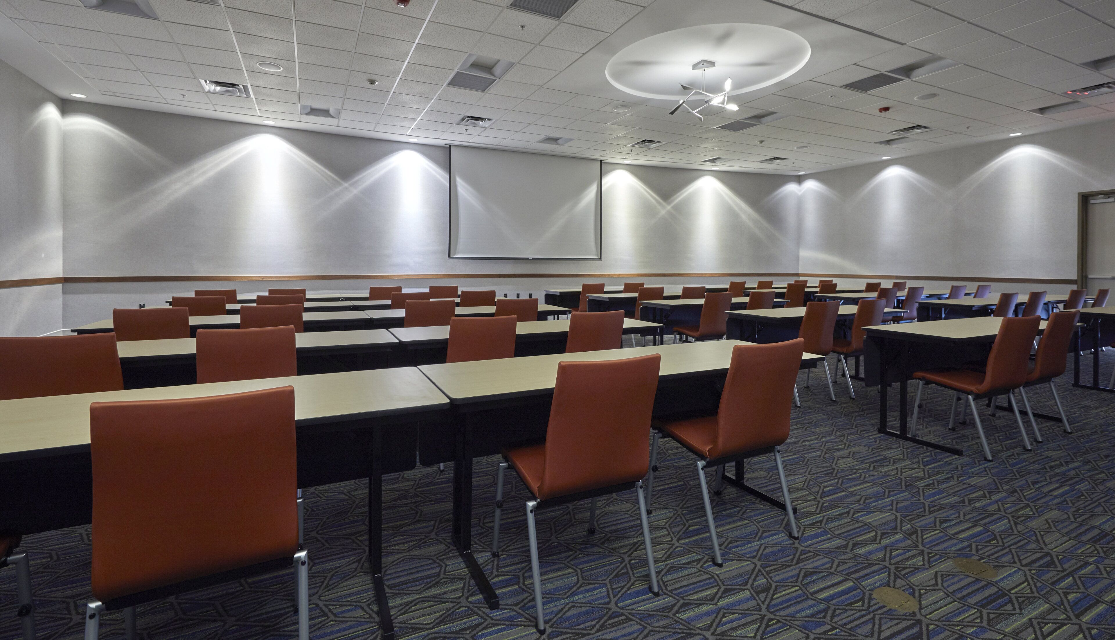 Une salle de conférence avec des rangées de tables et de chaises, un écran de projection et une moquette à motifs.
