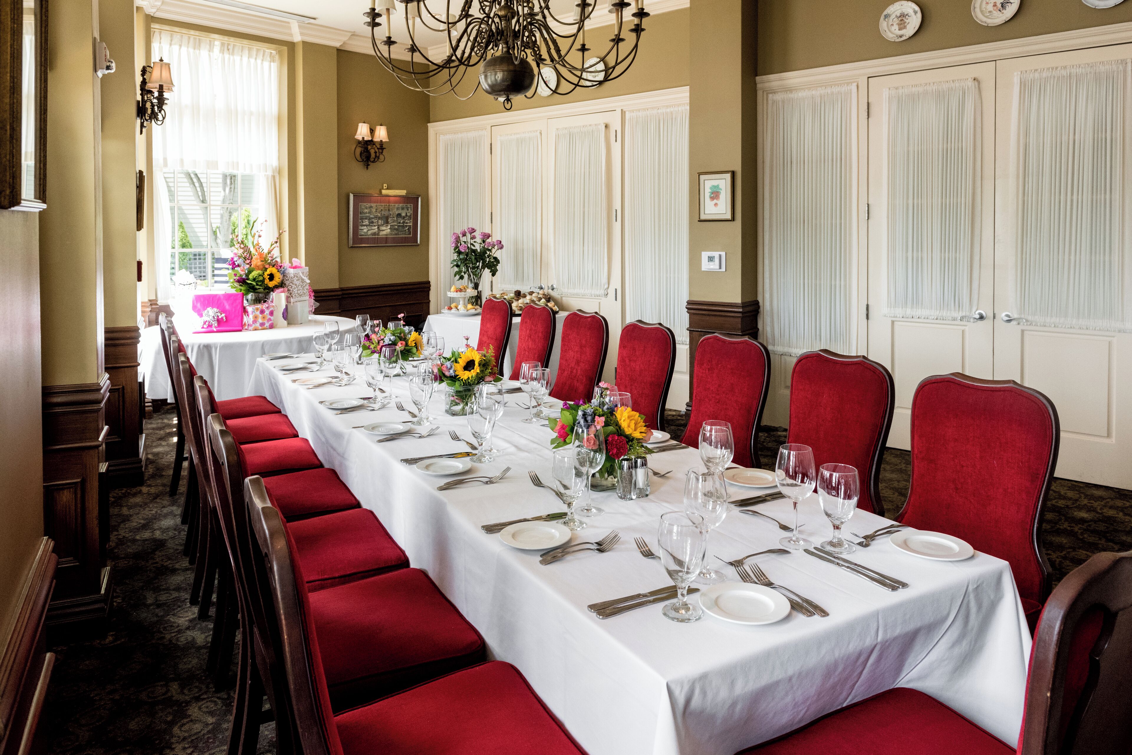 Una larga mesa de comedor dispuesta para una comida formal, con sillas tapizadas en rojo, manteles blancos y centros de mesa florales.