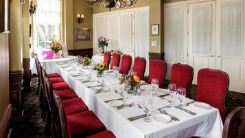 Una larga mesa de comedor dispuesta para una comida formal, con sillas tapizadas en rojo, manteles blancos y centros de mesa florales.