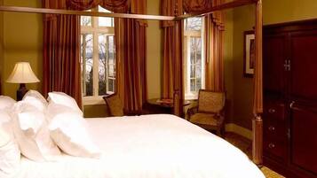 A four-poster bed with white linens, a bedside table with a lamp, a chair, and a wardrobe in a room with green walls and large windows.