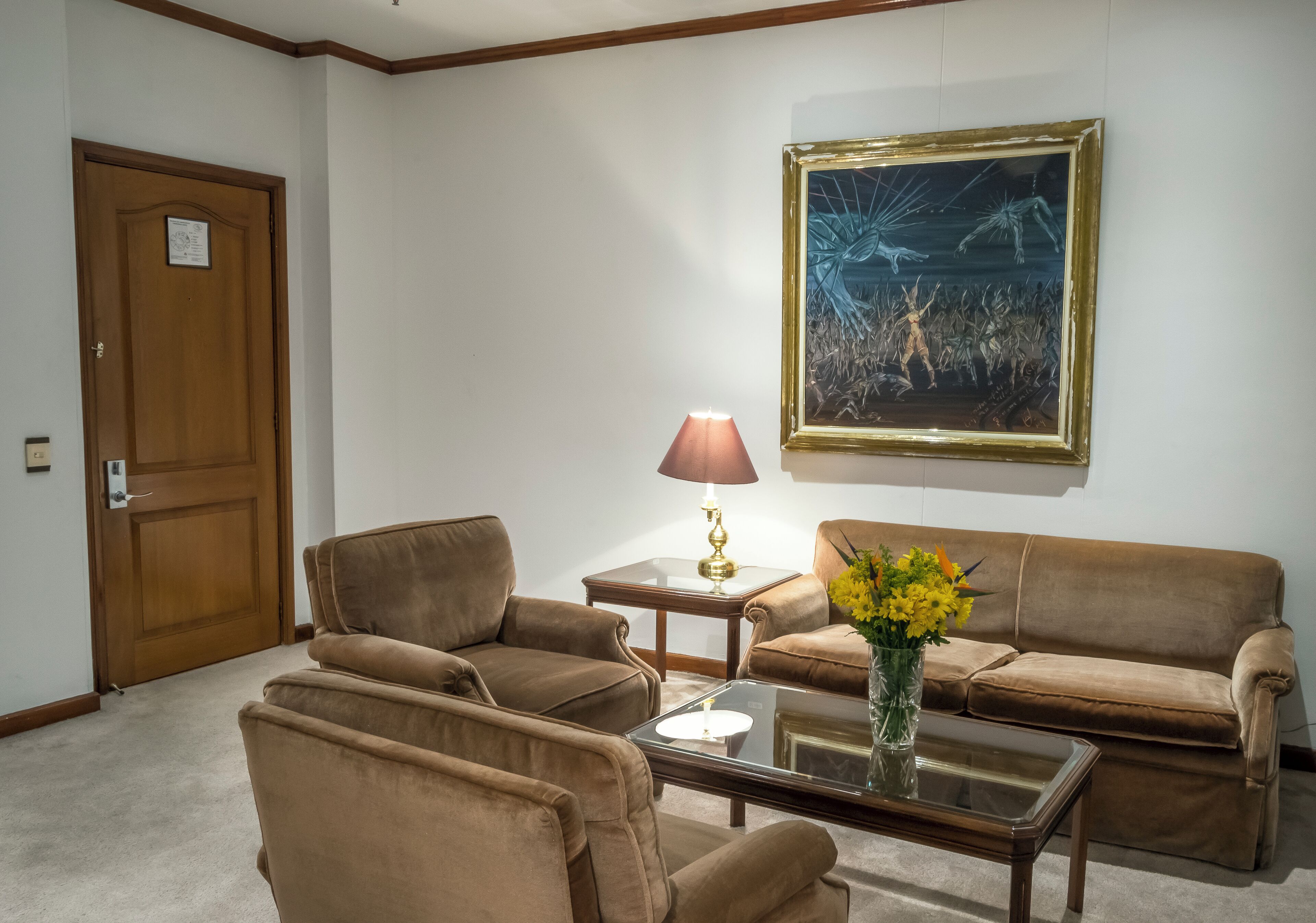 Una sala de estar con un sofá marrón, una mesa de centro de vidrio, una lámpara y una obra de arte enmarcada en la pared.