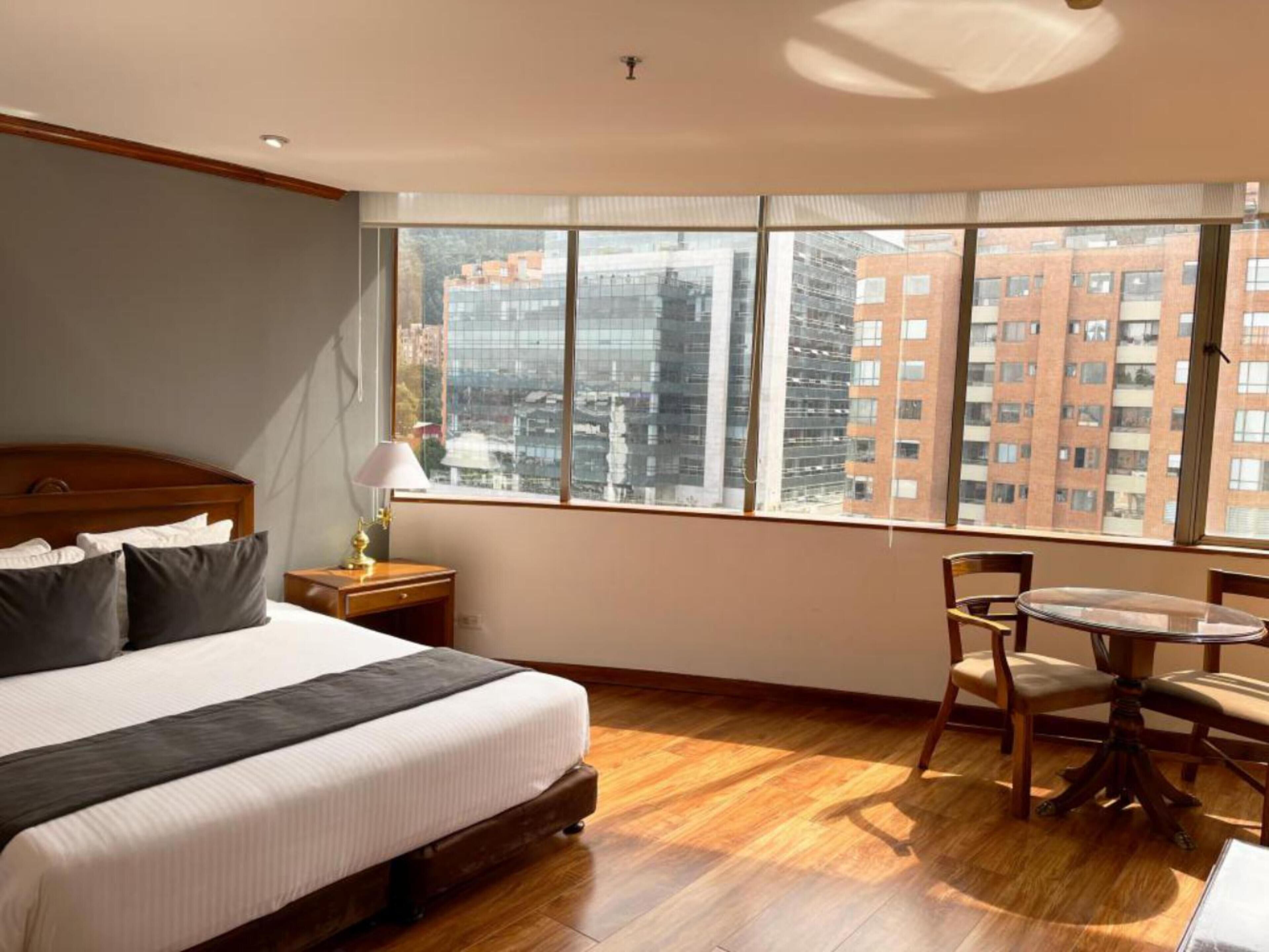 Habitación de hotel con una cama grande, una mesita de noche de madera, un escritorio con lámpara, una mesa redonda con sillas y un ventanal con vistas a la ciudad.