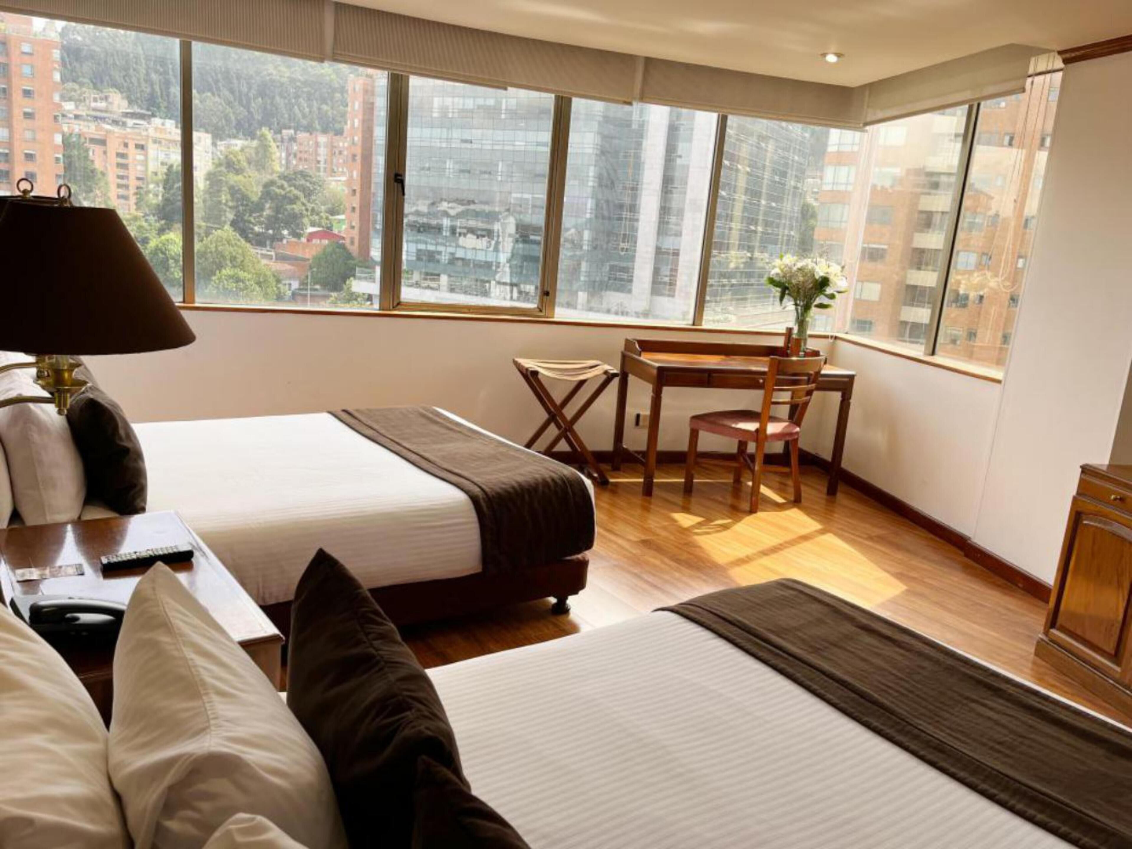 Habitación de hotel con cama, escritorio y silla. Se ve un ventanal con vistas a edificios y árboles.