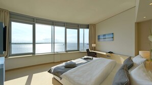 A modern hotel room with a large bed, a desk, and a view of the sea.