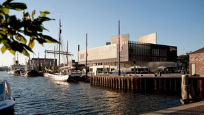A waterfront building with a modern design, a dock with boats, and a clear sky.