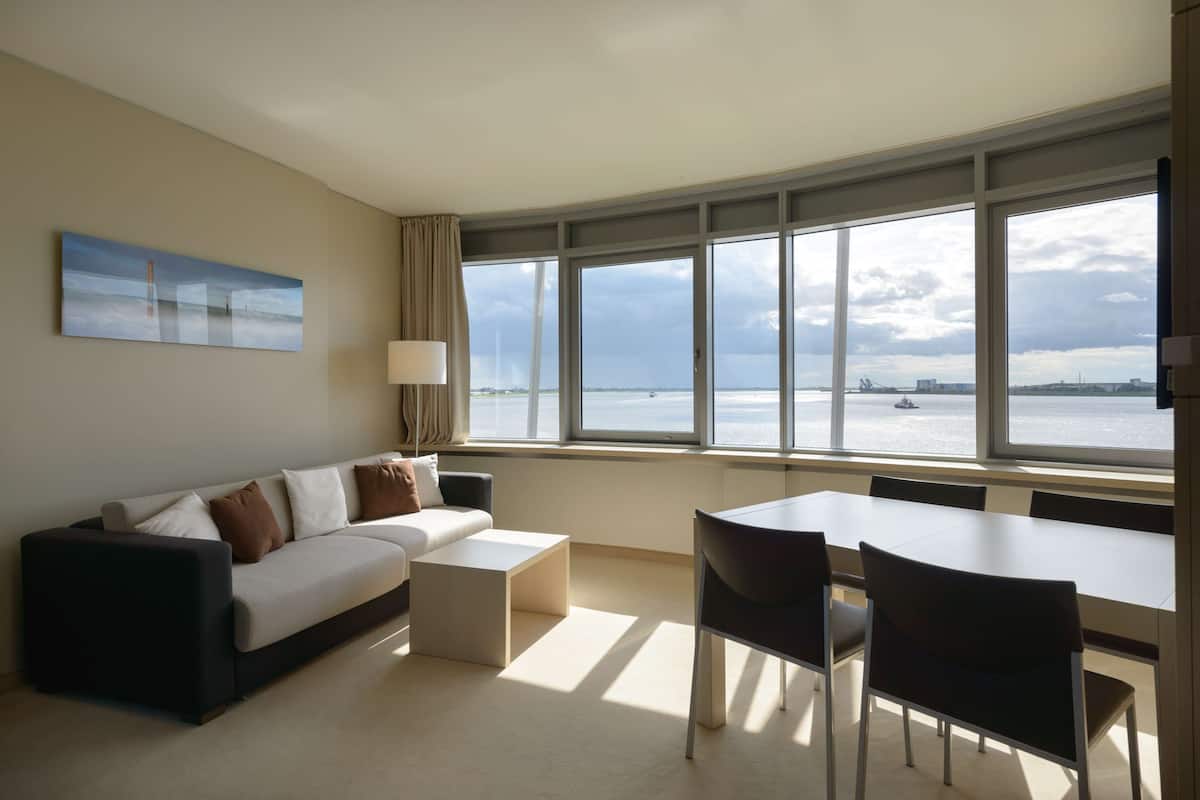 Een moderne hotelkamer met een groot raam met uitzicht op zee en een schip in de verte. Er is een bank, een klein tafeltje en een zithoek met stoelen.