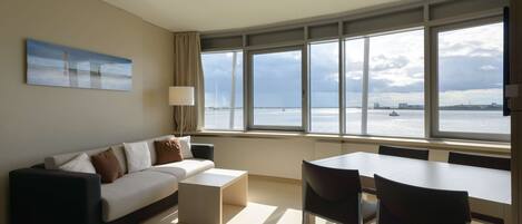 A modern hotel room with a large window offering a view of the sea and a distant ship. There is a sofa, a small table, and a dining area with chairs.