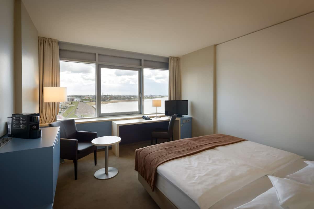 Een hotelkamer met een bed, bureau, stoel en uitzicht op de stad door grote ramen.