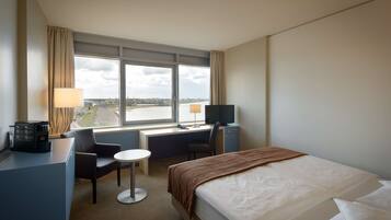 Ett hotellrum med en säng, ett skrivbord, en stol och utsikt över staden genom stora fönster.