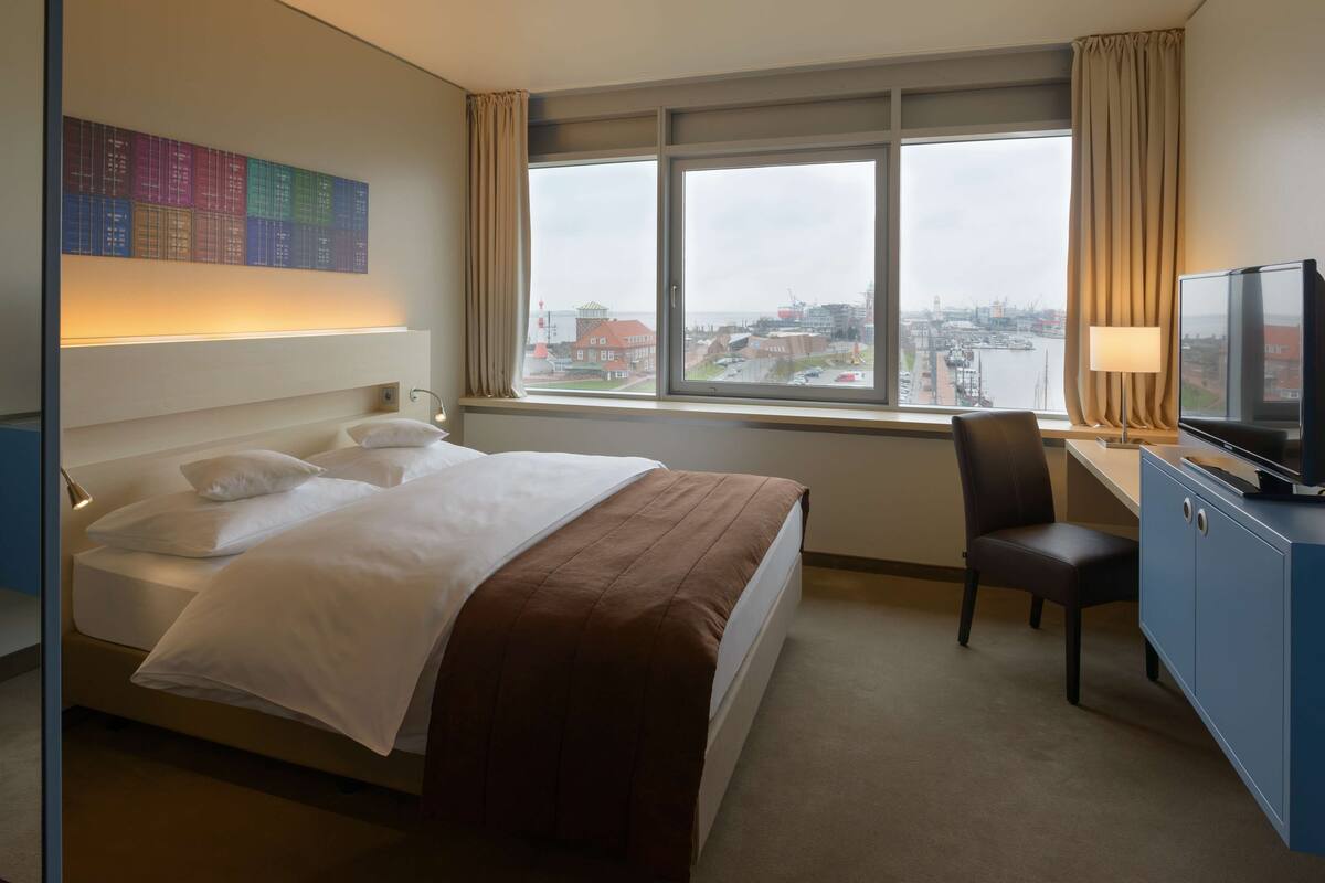 Hotelkamer met een bed, een bureau met stoel, een televisie en een groot raam met uitzicht op de stad.