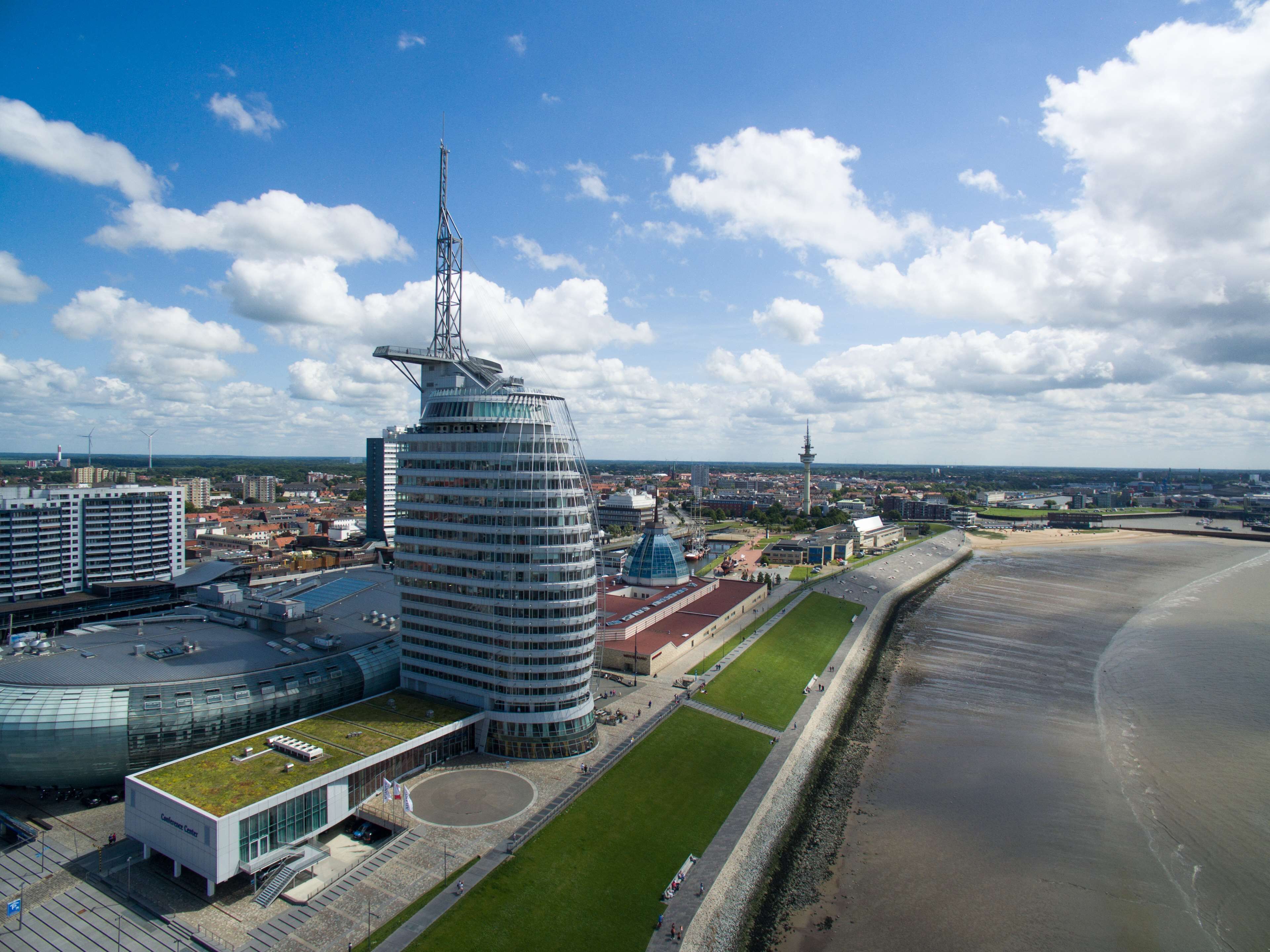 Um edifício moderno com um design curvo único, telhado verde e vista para a praia e paisagem urbana ao fundo.