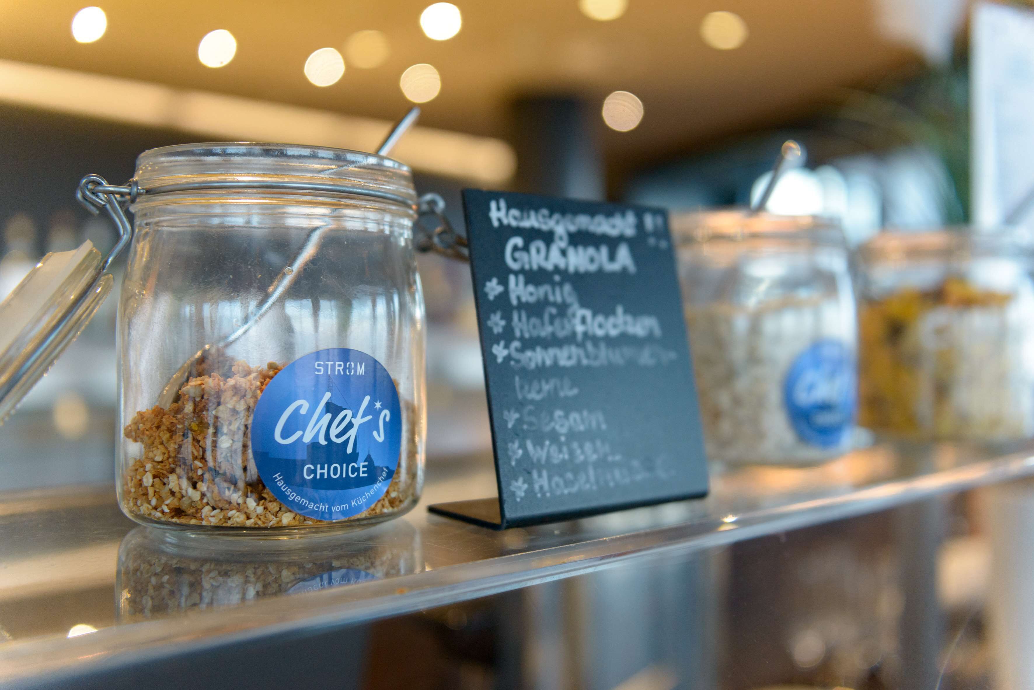 A glass jar labeled "Chef's Choice" with granola inside, placed on a shelf with a sign listing granola ingredients.