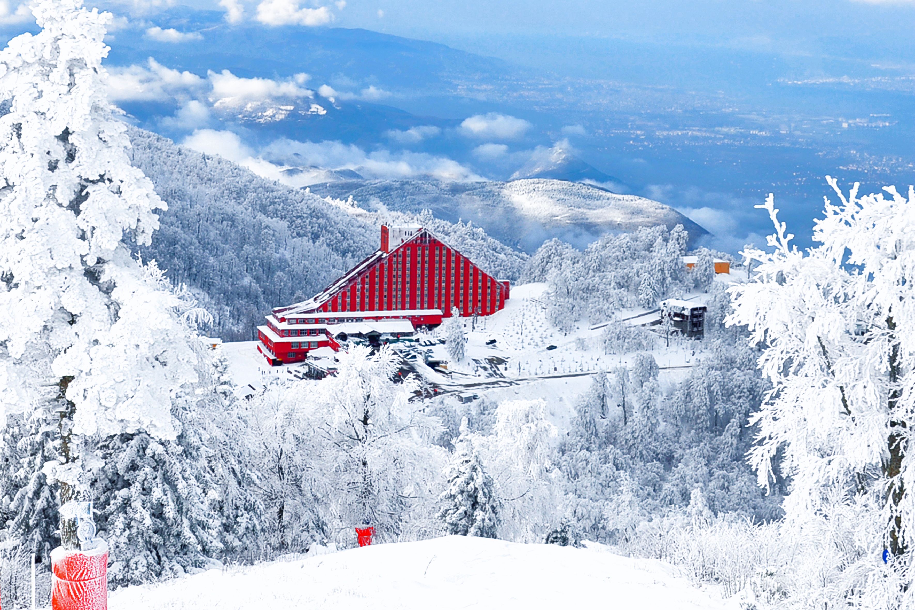 A snow-covered mountain landscape with a red building featuring a steep roof, surrounded by snow-laden trees and a clear sky.