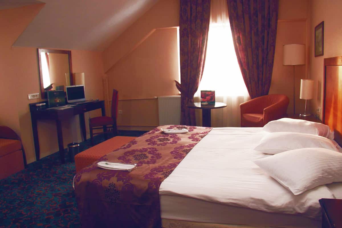 A hotel room with a bed, a desk with a laptop, a chair, a small table, and a lamp.