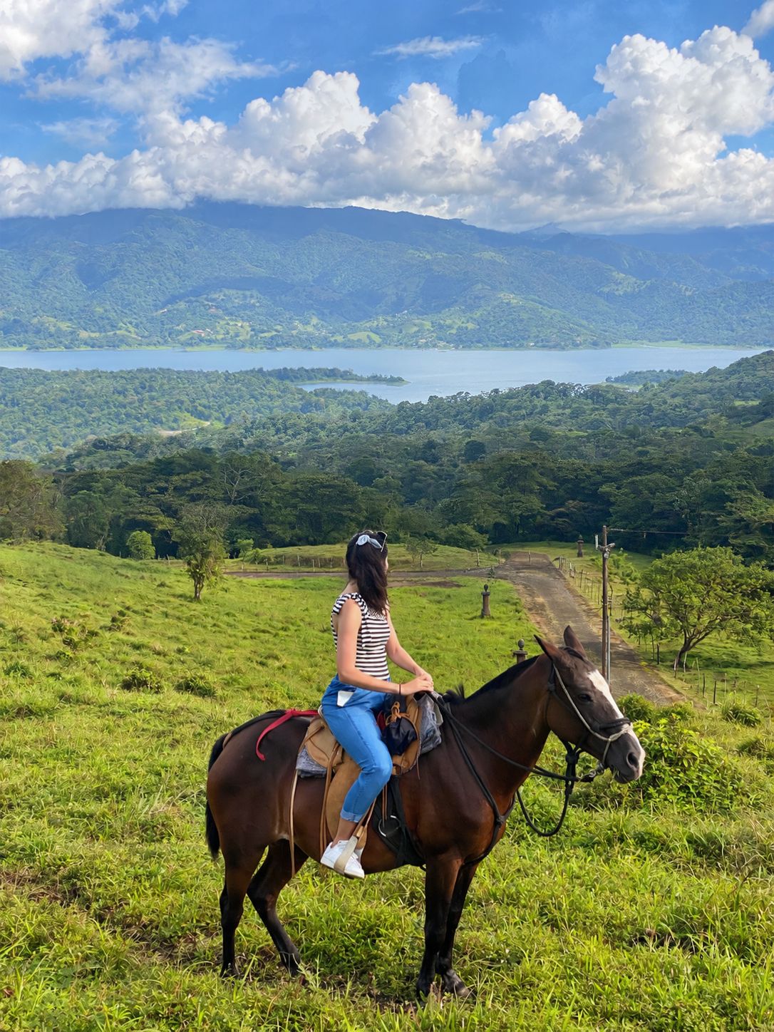 A person riding a horse on a grassy hill with a lake and mountains in the background.