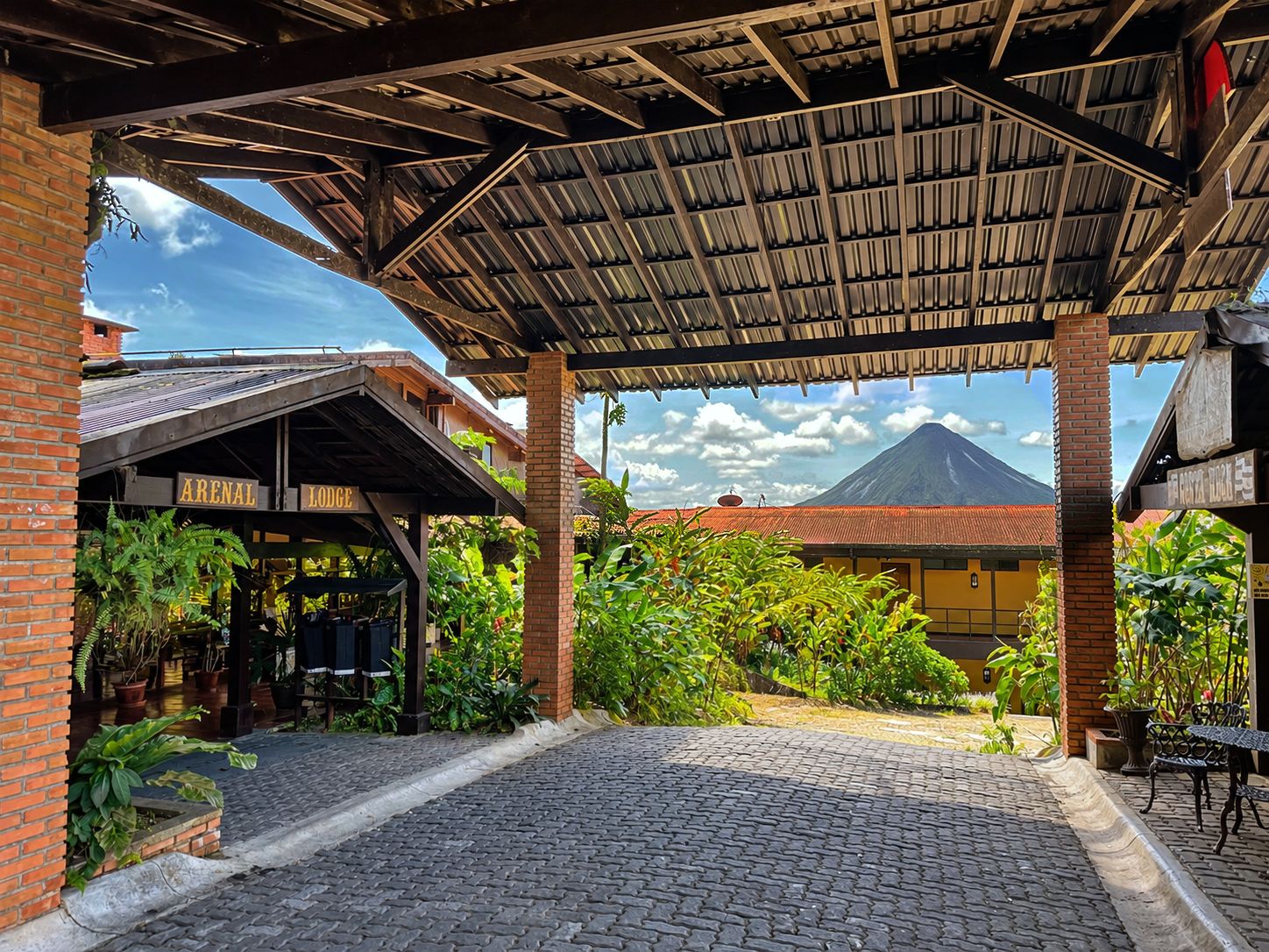 Un espacio al aire libre techado con piso de adoquines, un edificio de ladrillo con un letrero que dice 'ARENAL LODGE', y una vista de un volcán al fondo.