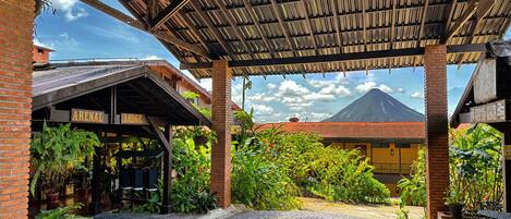 Un espacio al aire libre techado con piso de adoquines, un edificio de ladrillo con un letrero que dice 'ARENAL LODGE', y una vista de un volcán al fondo.