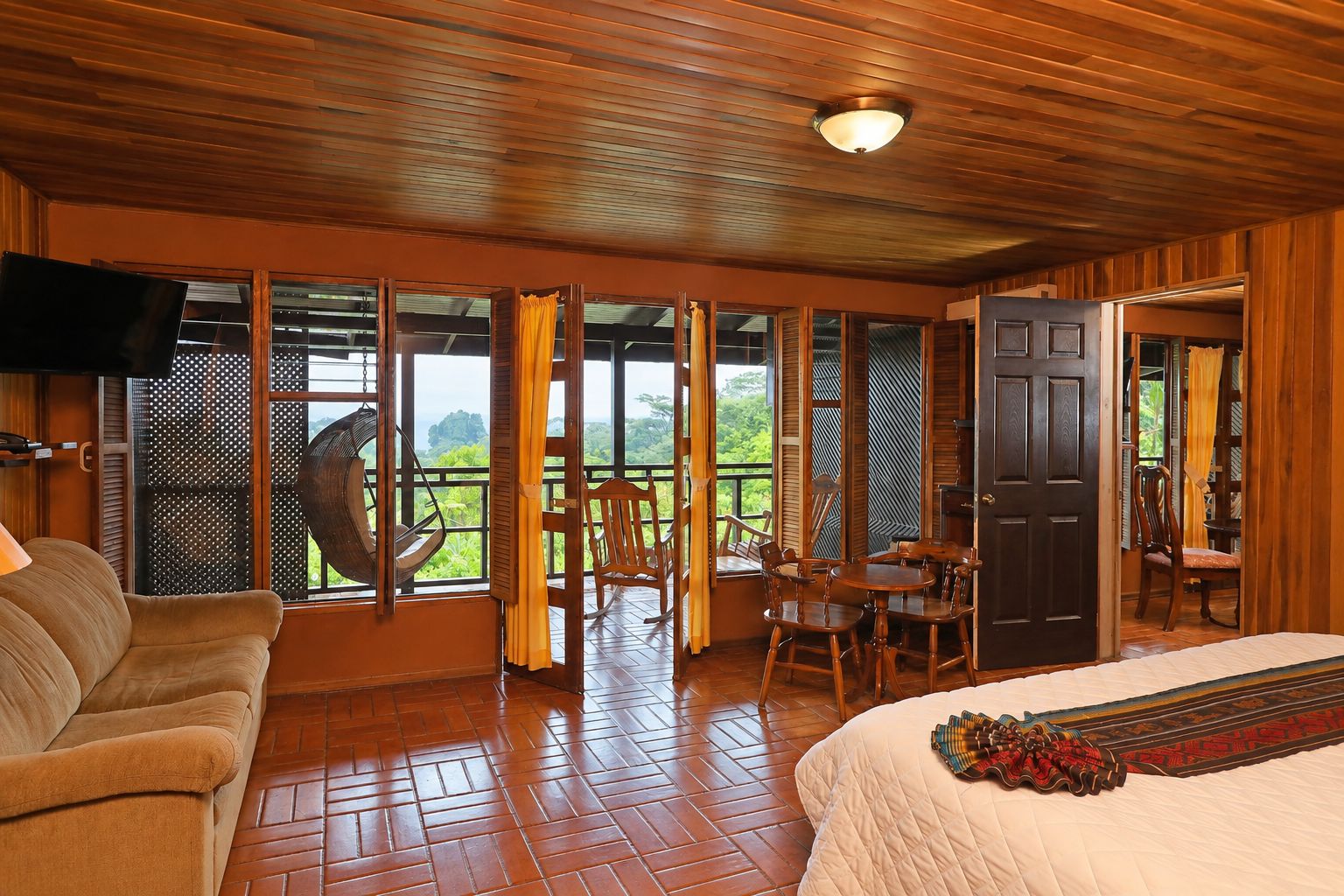 A room with a wooden ceiling, a bed with a decorative headboard, a sofa, a dining table with chairs, and a view through large windows.