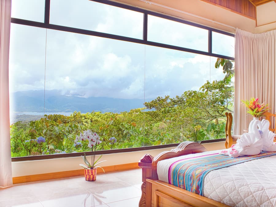 Una camera d'albergo con una grande finestra, un letto con una coperta colorata, un vaso con fiori e una testiera in legno.