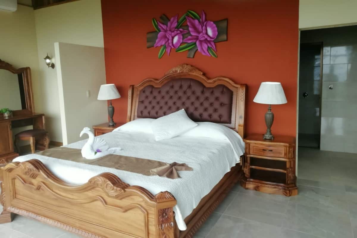 A large bed with a wooden headboard, two bedside tables with lamps, a floral artwork on the wall, and a mirror on the left.