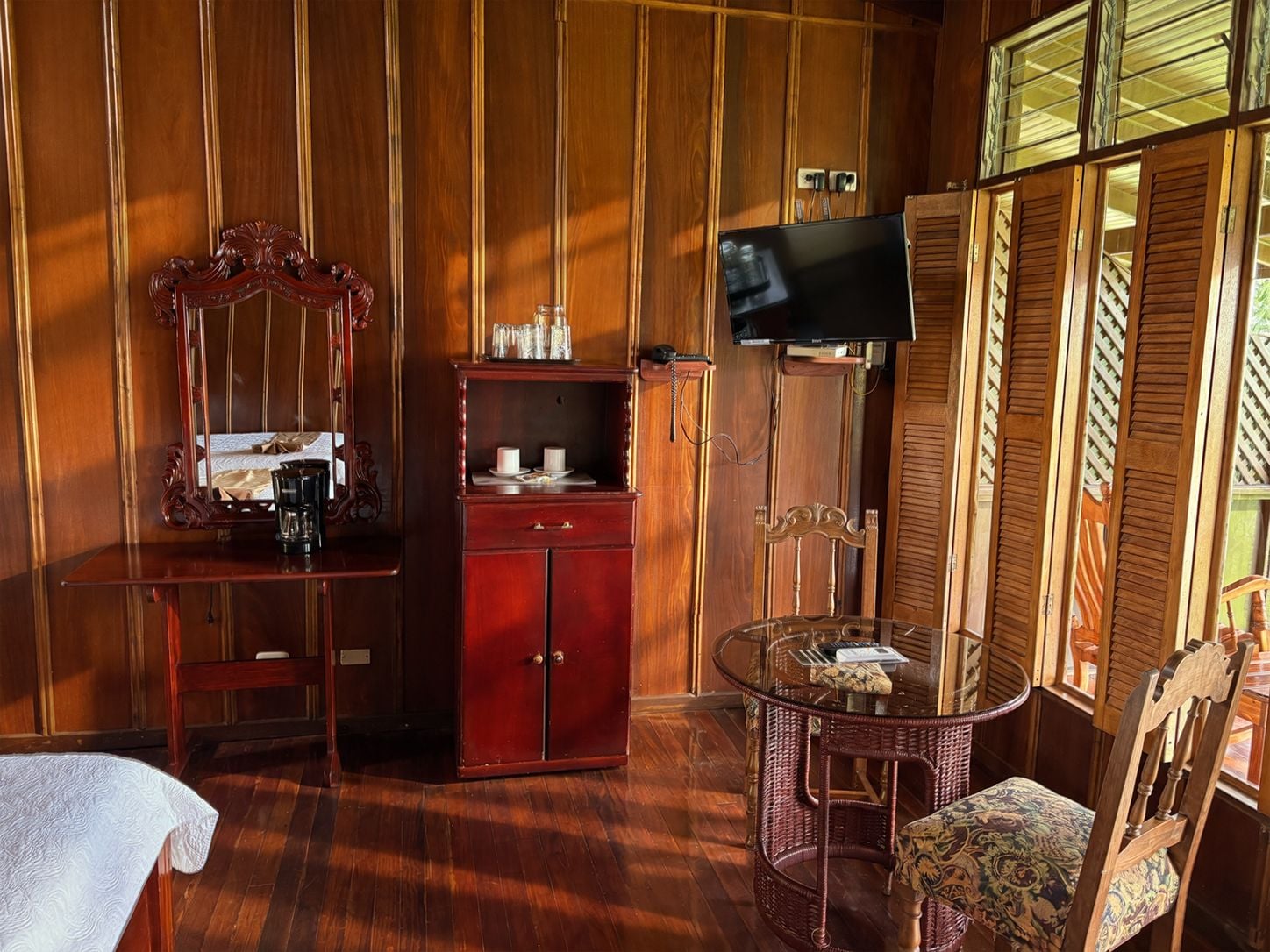 A room with a bed, a wooden wardrobe, a television, a glass table with a chair, and a window with wooden shutters.