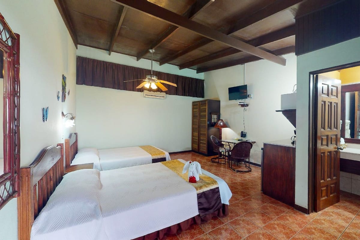 A room with two beds, a desk, a chair, a television, and a bathroom with a mirror.