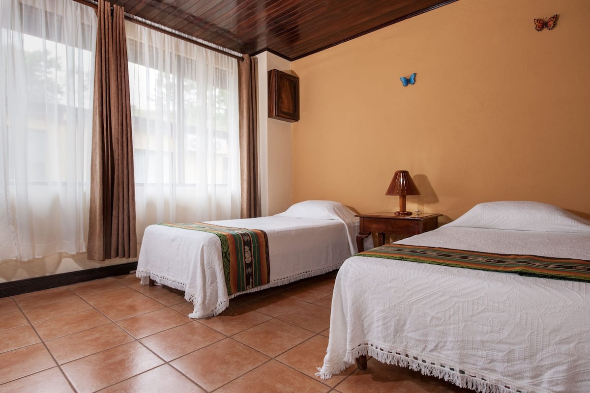 A room with two beds, a window with curtains, a small table with a lamp, and a butterfly decoration on the wall.