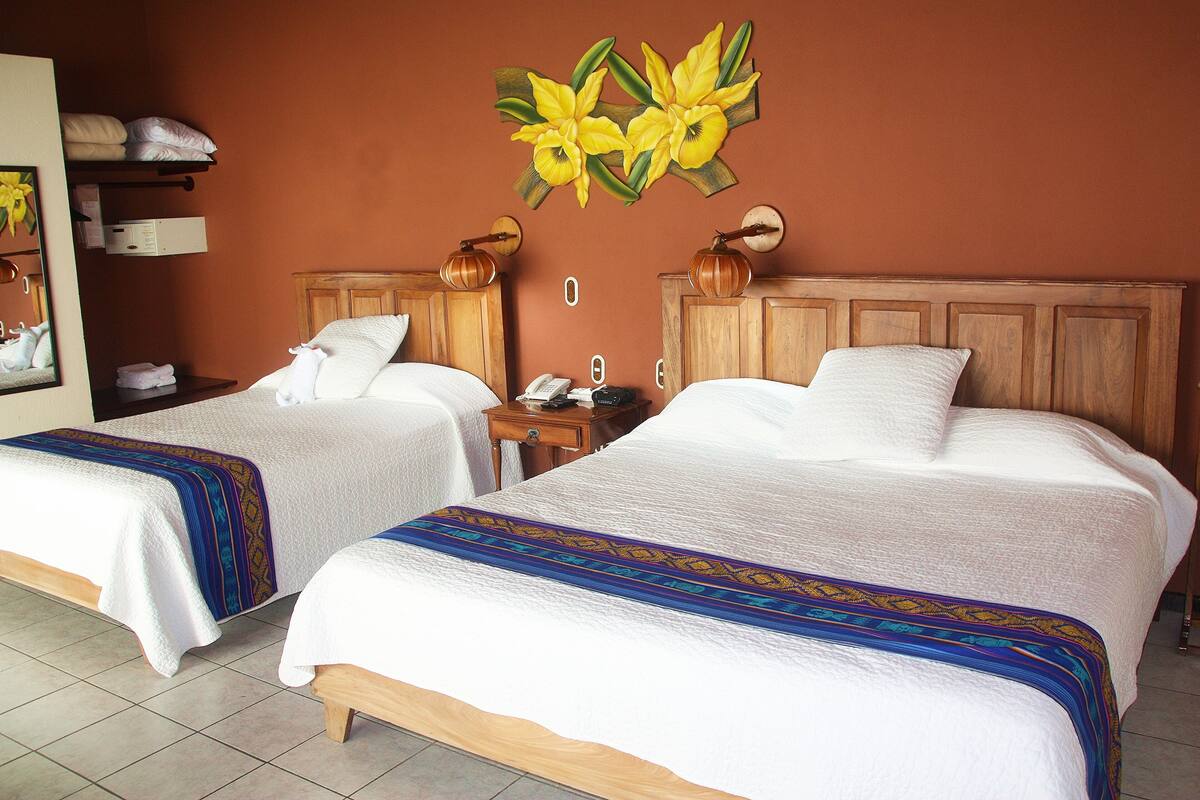 A hotel room with two beds, a wooden headboard, a nightstand with a phone, and a wall-mounted lamp.