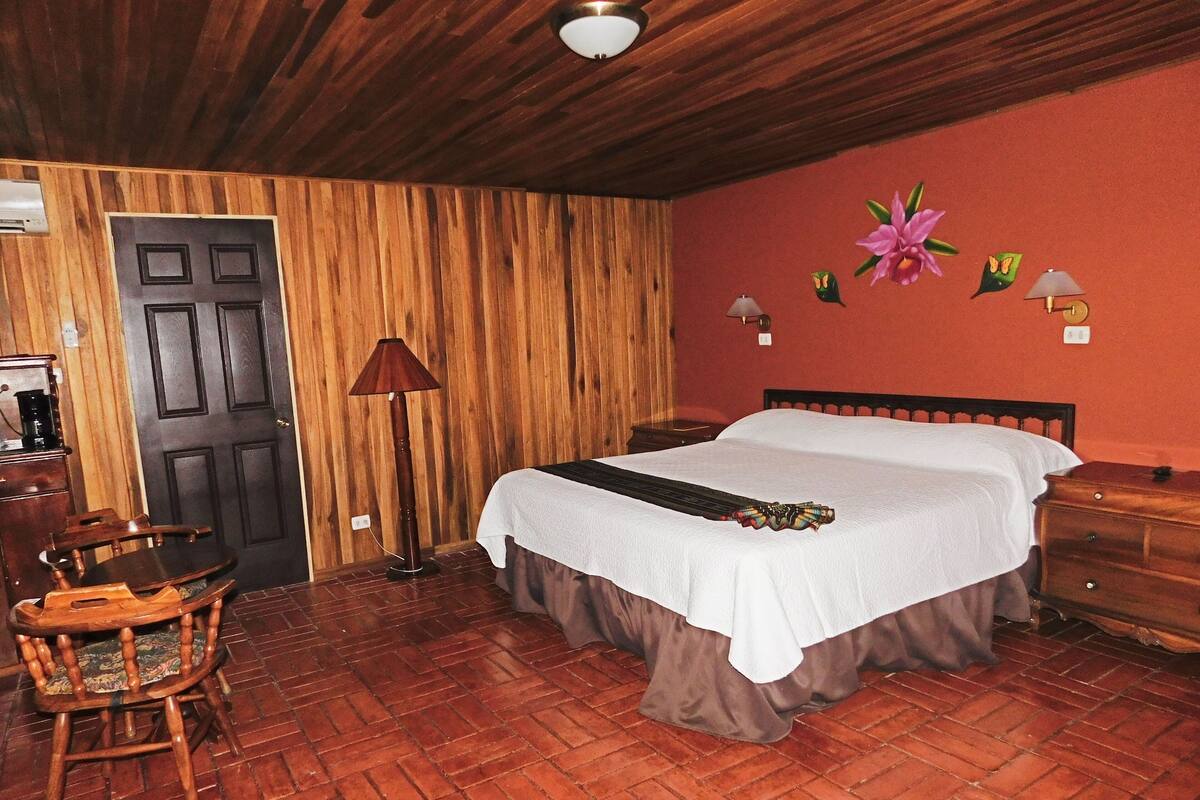 A room with a wooden floor, a bed with a white bedspread, a wooden chair, a desk with a lamp, and a door.