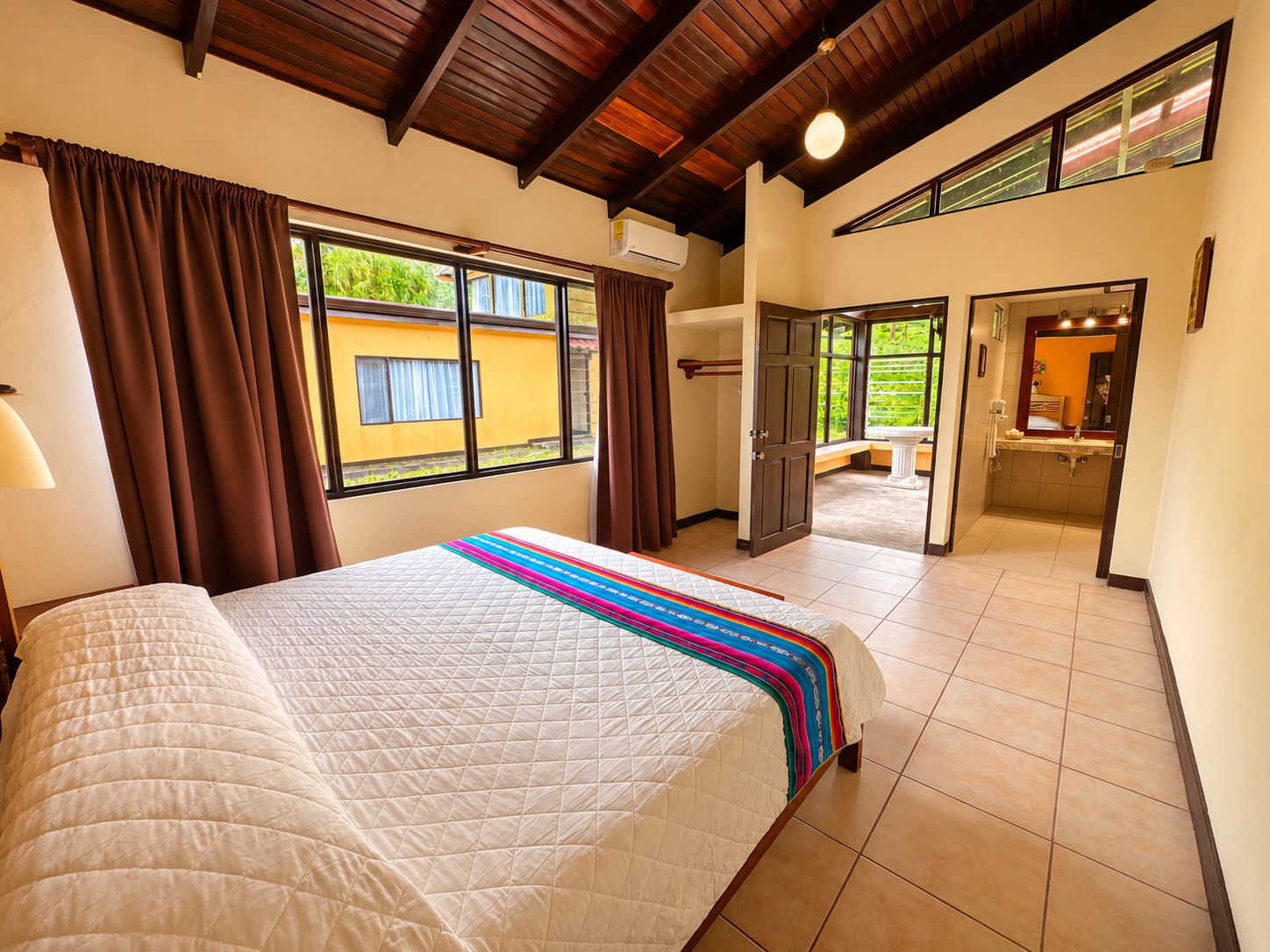 A hotel room with a large bed, a window with curtains, a bathroom with a sink and mirror, and a view of the outside.